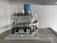 A complex mechanical apparatus with several metallic components, including pipes and cylinders, is prominently displayed. The machine features a blue cylindrical section at the top and is situated in a minimalist white room.