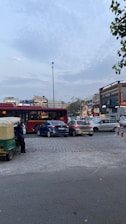 A busy urban street scene with several cars, a bus, and an auto rickshaw. A person stands next to the rickshaw. In the background, there are commercial buildings with illuminated signs and advertisements. The sky is partly cloudy, and trees are visible at the sides.