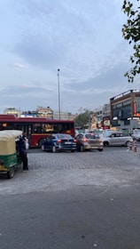 A busy urban street scene with several cars, a bus, and an auto rickshaw. A person stands next to the rickshaw. In the background, there are commercial buildings with illuminated signs and advertisements. The sky is partly cloudy, and trees are visible at the sides.