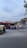A busy urban street scene with several cars, a bus, and an auto rickshaw. A person stands next to the rickshaw. In the background, there are commercial buildings with illuminated signs and advertisements. The sky is partly cloudy, and trees are visible at the sides.