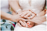 Several hands of different ages and skin tones are gently clasped together, symbolizing unity and support. The hands are adorned with various rings and bracelets, and some have colorful nail polish. The background includes a portion of a lace garment.
