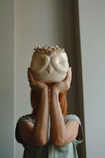 A person is holding an intricately designed ceramic vase shaped like an owl, in front of their face. The vase features detailed owl-like face imprints and decorative flowers on top. The background is a plain, neutral-colored wall.