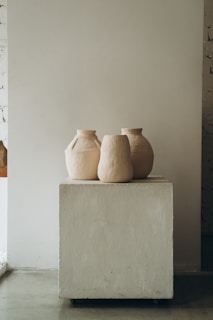 Three minimalist, neutral-toned ceramic vases are placed on a simple, square pedestal in a studio setting with an unfinished wall.