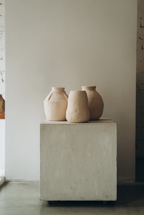 Three minimalist, neutral-toned ceramic vases are placed on a simple, square pedestal in a studio setting with an unfinished wall.