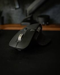 A sleek black wireless computer mouse with a metallic scroll wheel and ergonomic design placed on a dark surface. The background is out of focus, featuring a blurred tech environment.