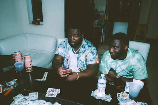 Two individuals are sitting at a table engaged in a card game. Various playing cards, poker chips, and items are scattered on the table. Arizona tea cans and drinks are present, and both people appear relaxed and focused.