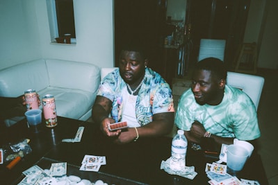 Two individuals are sitting at a table engaged in a card game. Various playing cards, poker chips, and items are scattered on the table. Arizona tea cans and drinks are present, and both people appear relaxed and focused.
