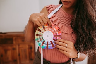 A person wearing a pink knitted top holds a color wheel with an array of vibrant color swatches arranged in a circular pattern. The person wears a ring and a watch on their left hand. A measuring tape drapes around their neck, and there is a wooden piece of furniture in the background.