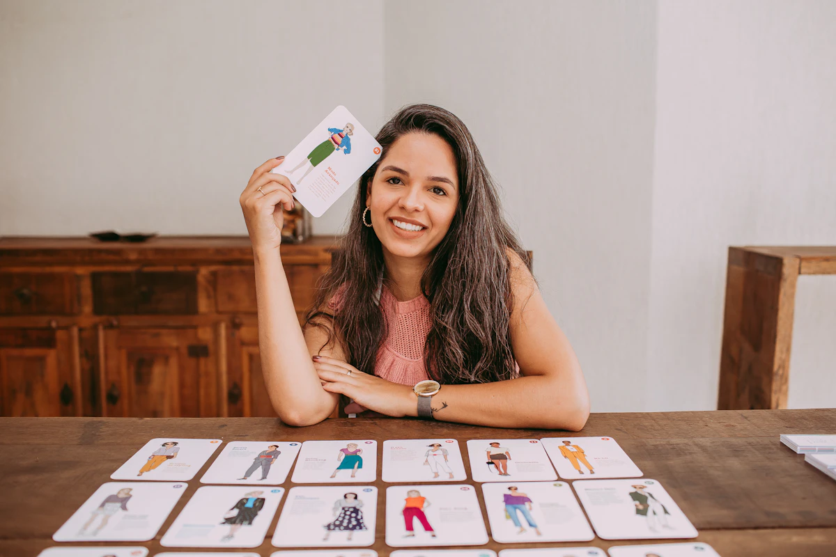 Une femme souriante assise à une table en bois lève une carte illustrée, plusieurs rangées de cartes du même jeu étalées devant elle