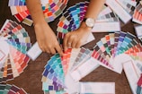 A variety of color swatches fan out across a wooden surface. Two hands point to specific swatches, with one hand wearing a watch. The swatches cover a wide spectrum of colors, including blues, greens, reds, and neutrals.