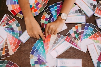 A variety of color swatches fan out across a wooden surface. Two hands point to specific swatches, with one hand wearing a watch. The swatches cover a wide spectrum of colors, including blues, greens, reds, and neutrals.