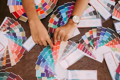 A variety of color swatches fan out across a wooden surface. Two hands point to specific swatches, with one hand wearing a watch. The swatches cover a wide spectrum of colors, including blues, greens, reds, and neutrals.