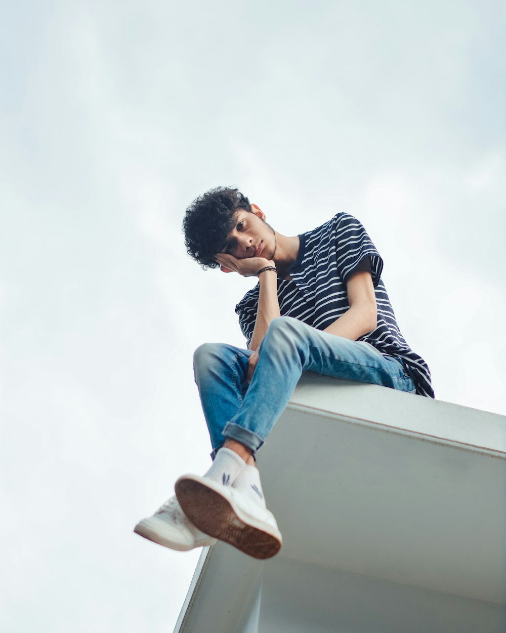 Homme en jean bleu et chemise rayée blanche et noire assis sur un mur de béton blanc