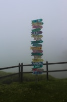 A colorful signpost stands in a foggy landscape, displaying arrows pointing in different directions with names of countries written on them. The scene is surrounded by a wooden fence and lush green grass, adding to the serene and mystical atmosphere.