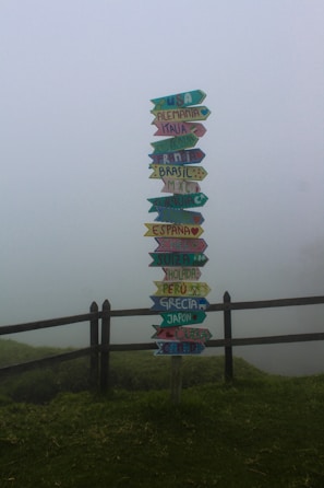 A colorful signpost stands in a foggy landscape, displaying arrows pointing in different directions with names of countries written on them. The scene is surrounded by a wooden fence and lush green grass, adding to the serene and mystical atmosphere.