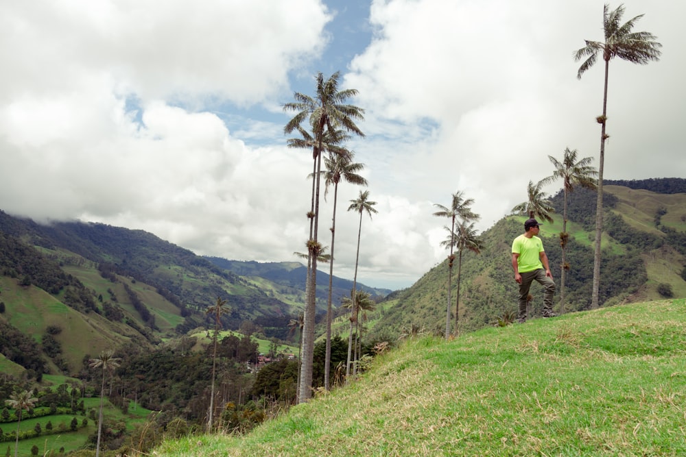 man in yellow shirt standing on green grass field near green mountain under white cloudy sky