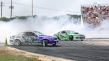 Two cars are drifting on a racetrack surrounded by a cloud of smoke. There is a crowd of spectators seated on bleachers in the background, watching the thrilling event. The cars are adorned with colorful decals, one predominantly purple and the other green.