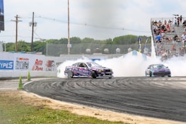 A vibrant car race scene with two cars drifting around a track, producing thick white smoke. The cars, adorned with colorful graphics, add energy to the setting, while a crowd of spectators watches intently from bleachers. The surrounding environment features green foliage and promotional banners on the boundary walls.