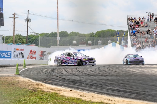 A vibrant car race scene with two cars drifting around a track, producing thick white smoke. The cars, adorned with colorful graphics, add energy to the setting, while a crowd of spectators watches intently from bleachers. The surrounding environment features green foliage and promotional banners on the boundary walls.