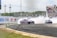A vibrant car race scene with two cars drifting around a track, producing thick white smoke. The cars, adorned with colorful graphics, add energy to the setting, while a crowd of spectators watches intently from bleachers. The surrounding environment features green foliage and promotional banners on the boundary walls.