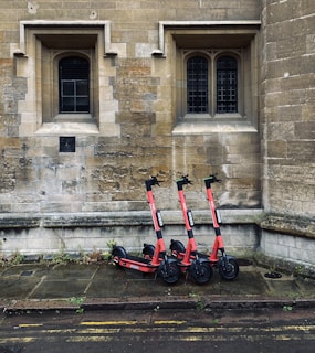 Three red electric scooters are parked on a stone-paved sidewalk in front of an old stone building with two arched windows.