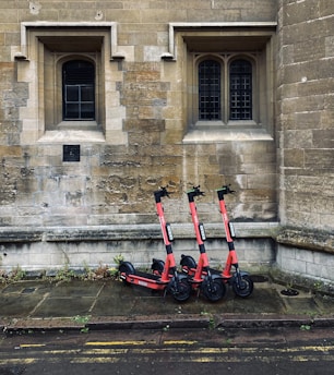 Three red electric scooters are parked on a stone-paved sidewalk in front of an old stone building with two arched windows.