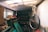 A cluttered, dimly lit storage room filled with various objects. A bicycle is leaning against the wall on the right, and the ceiling appears to have gaps, letting some light in. Shelves are overloaded with boxes, cans, and other items. Dust and signs of neglect are evident throughout the space.