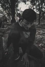 A figure crouches in a wooded area, wearing a dark hoodie and a Guy Fawkes mask, with smoke surrounding them. The setting appears to be dimly lit, with trees and natural elements forming the background.