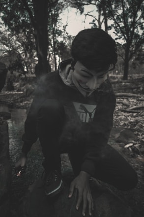 A figure crouches in a wooded area, wearing a dark hoodie and a Guy Fawkes mask, with smoke surrounding them. The setting appears to be dimly lit, with trees and natural elements forming the background.