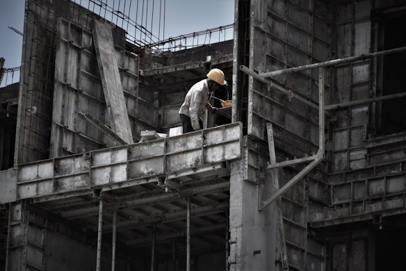 A worker wearing a yellow hard hat is positioned on the edge of a partially constructed building, which features exposed concrete forms and metal reinforcements. The surroundings convey an industrial and somewhat rugged atmosphere.