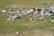 A field scattered with litter, including plastic bottles, cardboard boxes, and various other trash items. The grass is patchy and mixed with dry and green areas, and the amount of waste suggests a lack of proper disposal or care for the environment.