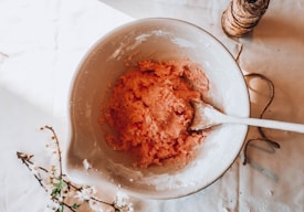 A ceramic mixing bowl contains a pink dough mixture and is accompanied by a wooden spoon. The scene is set on a white tablecloth with twine and delicate white blossoms scattered around.