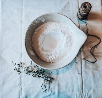 A bowl filled with a fine white powder resembling flour sits on a lightly wrinkled, pale surface. Next to the bowl, a coil of twine is partially unraveled, and delicate flowers rest on the surface, bringing a touch of nature to the setting.