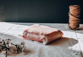 A neatly wrapped package sits on a light-colored tablecloth. Next to it is a spool of brown twine, and some delicate white flowers scattered in the foreground. The scene is softly lit with shadows creating a calm atmosphere.