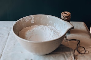 A large ceramic mixing bowl filled with white flour sits on a tablecloth-covered surface. Next to the bowl is a ball of twine with a loose end trailing off towards the bottom right corner.
