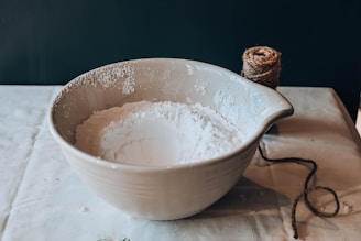A large ceramic mixing bowl filled with white flour sits on a tablecloth-covered surface. Next to the bowl is a ball of twine with a loose end trailing off towards the bottom right corner.