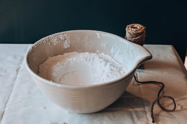 A large ceramic mixing bowl filled with white flour sits on a tablecloth-covered surface. Next to the bowl is a ball of twine with a loose end trailing off towards the bottom right corner.