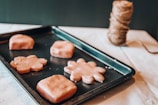 A baking tray holds several pieces of dough shaped like flowers and squares, resting on a light-colored surface. A spool of thick twine is visible in the background, partially out of focus.
