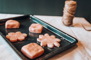 A baking tray holds several pieces of dough shaped like flowers and squares, resting on a light-colored surface. A spool of thick twine is visible in the background, partially out of focus.