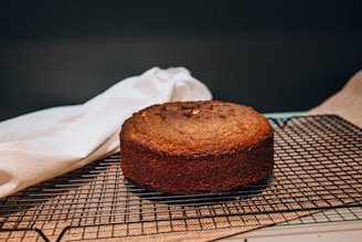 A freshly baked brown cake resting on a black cooling rack. A white cloth is draped next to it, while the background is softly focused, accentuating the baked item. The surface appears slightly textured, suggesting a classic homemade cake.