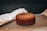 A freshly baked brown cake resting on a black cooling rack. A white cloth is draped next to it, while the background is softly focused, accentuating the baked item. The surface appears slightly textured, suggesting a classic homemade cake.