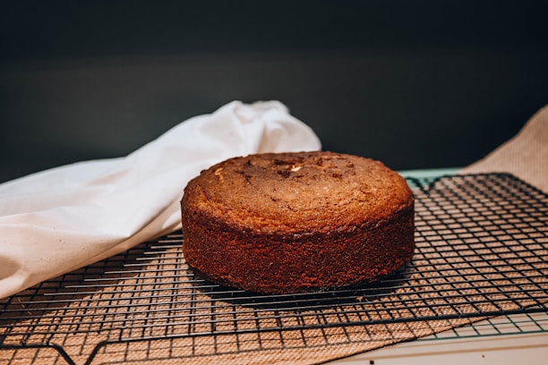 A freshly baked brown cake resting on a black cooling rack. A white cloth is draped next to it, while the background is softly focused, accentuating the baked item. The surface appears slightly textured, suggesting a classic homemade cake.