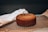 A freshly baked brown cake resting on a black cooling rack. A white cloth is draped next to it, while the background is softly focused, accentuating the baked item. The surface appears slightly textured, suggesting a classic homemade cake.