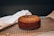 A freshly baked brown cake resting on a black cooling rack. A white cloth is draped next to it, while the background is softly focused, accentuating the baked item. The surface appears slightly textured, suggesting a classic homemade cake.