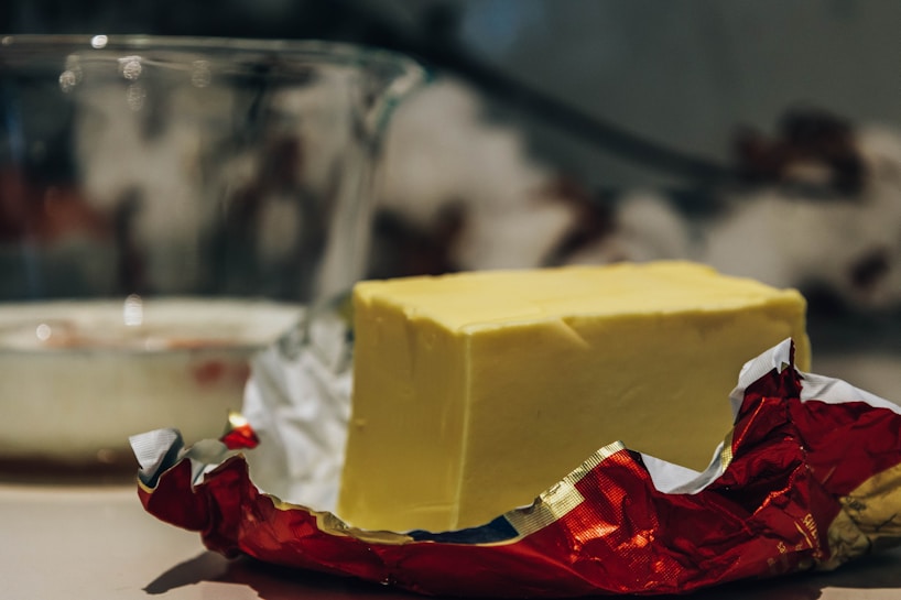 A block of butter is placed on a surface, partially unwrapped from its red and silver foil packaging. In the background, there is a transparent glass bowl with a blurred interior, suggesting a kitchen setting.