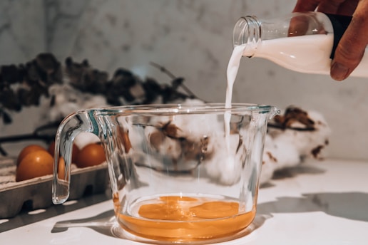 A clear glass measuring cup contains broken egg yolks, with milk being poured from a bottle into the cup. The background includes brown eggs in a carton and dried flowers.