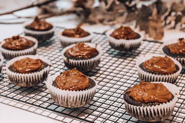Several chocolate cupcakes topped with a rich, brown frosting are arranged on a cooling rack. Each cupcake sits in a white paper liner, and the focus highlights the creamy texture of the frosting. The background is slightly blurred, emphasizing the cupcakes as the main subject.