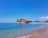 A picturesque coastal scene featuring a historic island with stone buildings topped by terracotta roofs. The island is surrounded by clear blue waters, and the distant mountains create a serene backdrop. People are enjoying the beach and swimming in the calm sea.