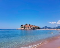 A picturesque coastal scene featuring a historic island with stone buildings topped by terracotta roofs. The island is surrounded by clear blue waters, and the distant mountains create a serene backdrop. People are enjoying the beach and swimming in the calm sea.