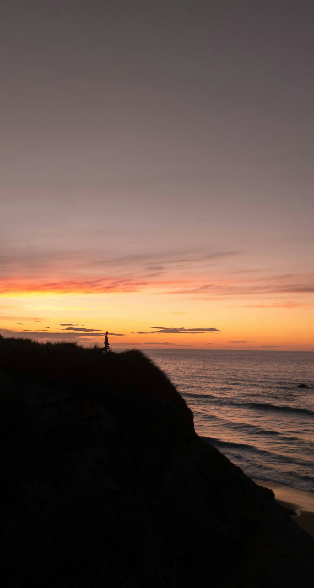 silhouette of person standing on rock formation near body of water during sunset